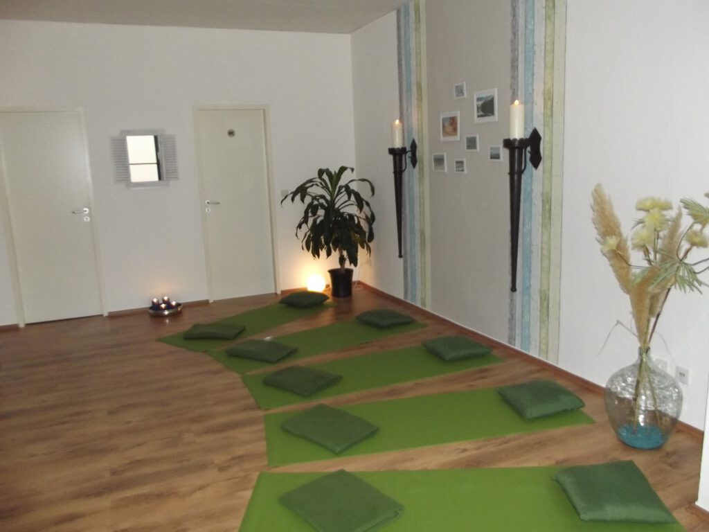 Hier abgebildet ist ein Entspannungsraum mit ausgelegten grünen Yogamatten. Auf den Yogamatten liegen jeweils Decken und Kissen. Das Licht des Raums angenehm warm durch Kerzenschein.
